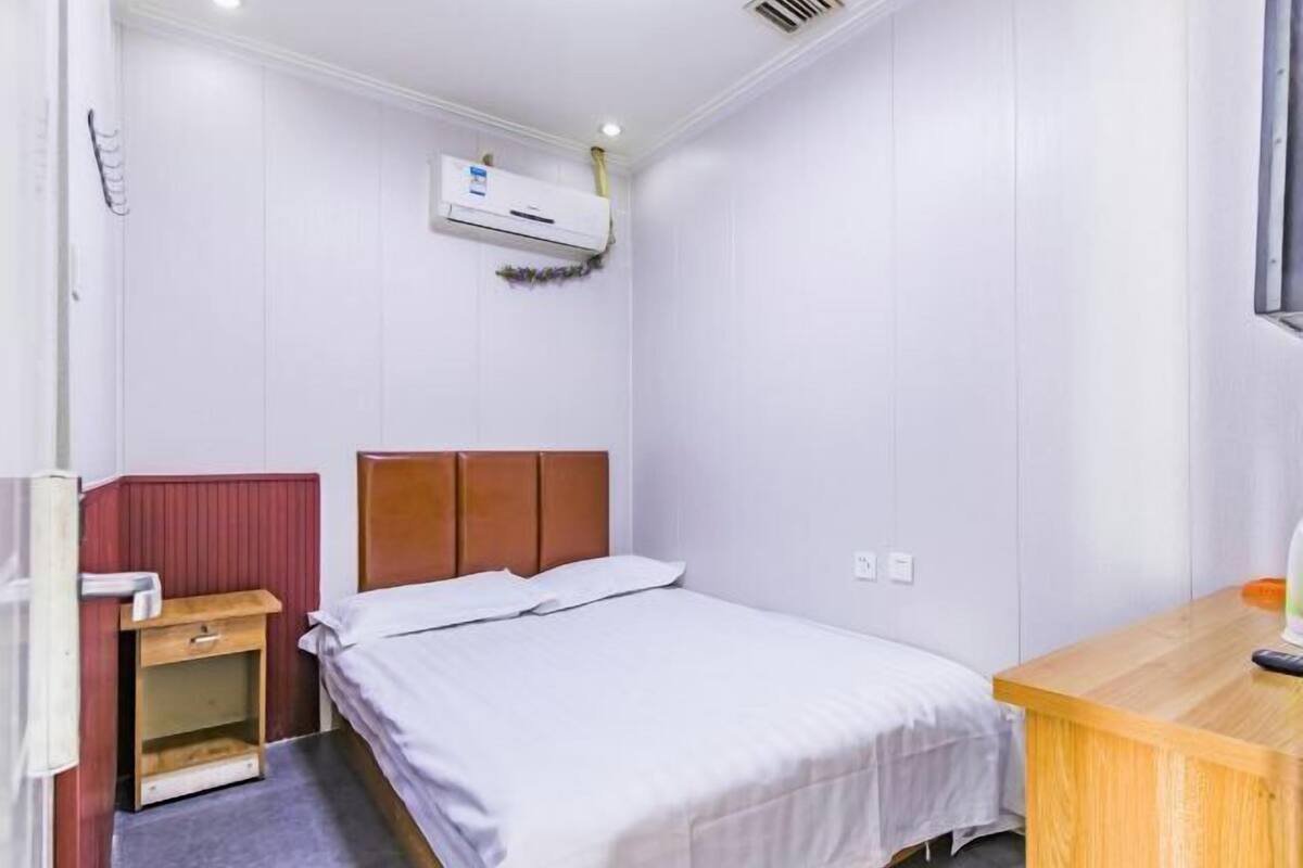 Una habitación pequeña con una cama individual, cabecera de madera, mesita de noche de madera, escritorio y un equipo de aire acondicionado montado en la pared.