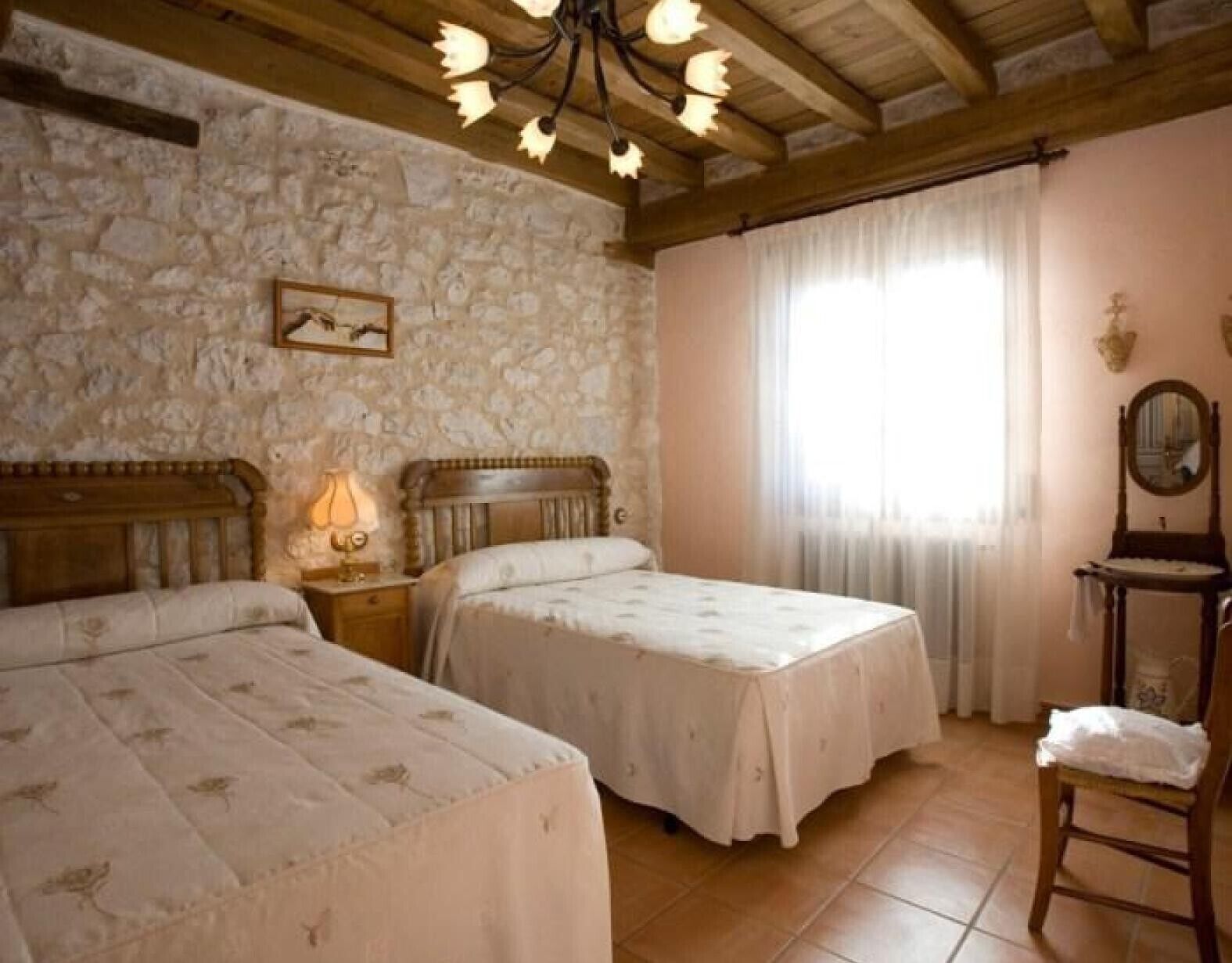 Ein Schlafzimmer mit zwei Betten, einer Steinwand, einer Holzbalkendecke, einem Kronleuchter, einem Nachttisch, einem Spiegel und einem Fenster mit Vorhängen.