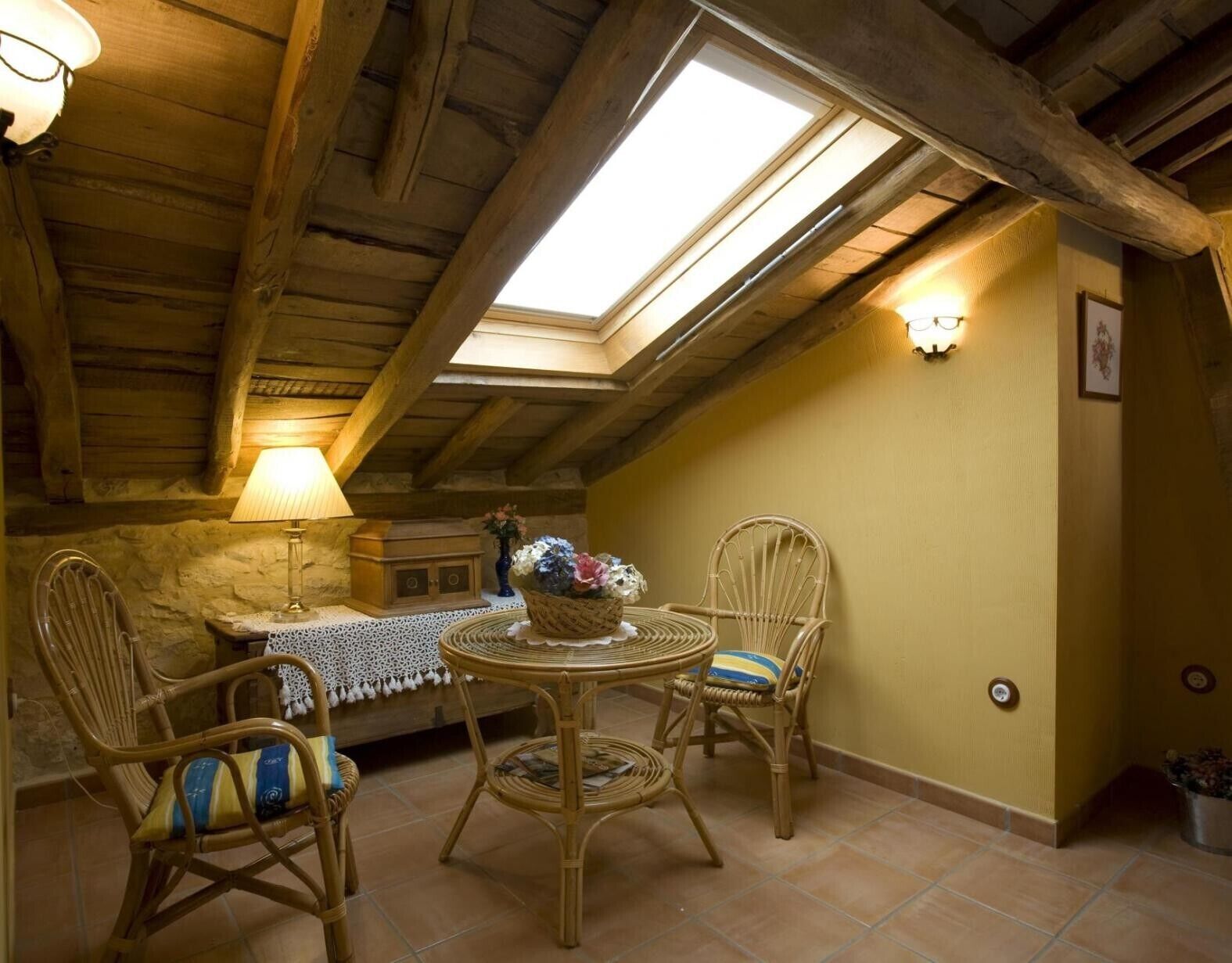 A cozy room with a wooden ceiling, a round table with chairs, a lamp, and a vase with flowers.
