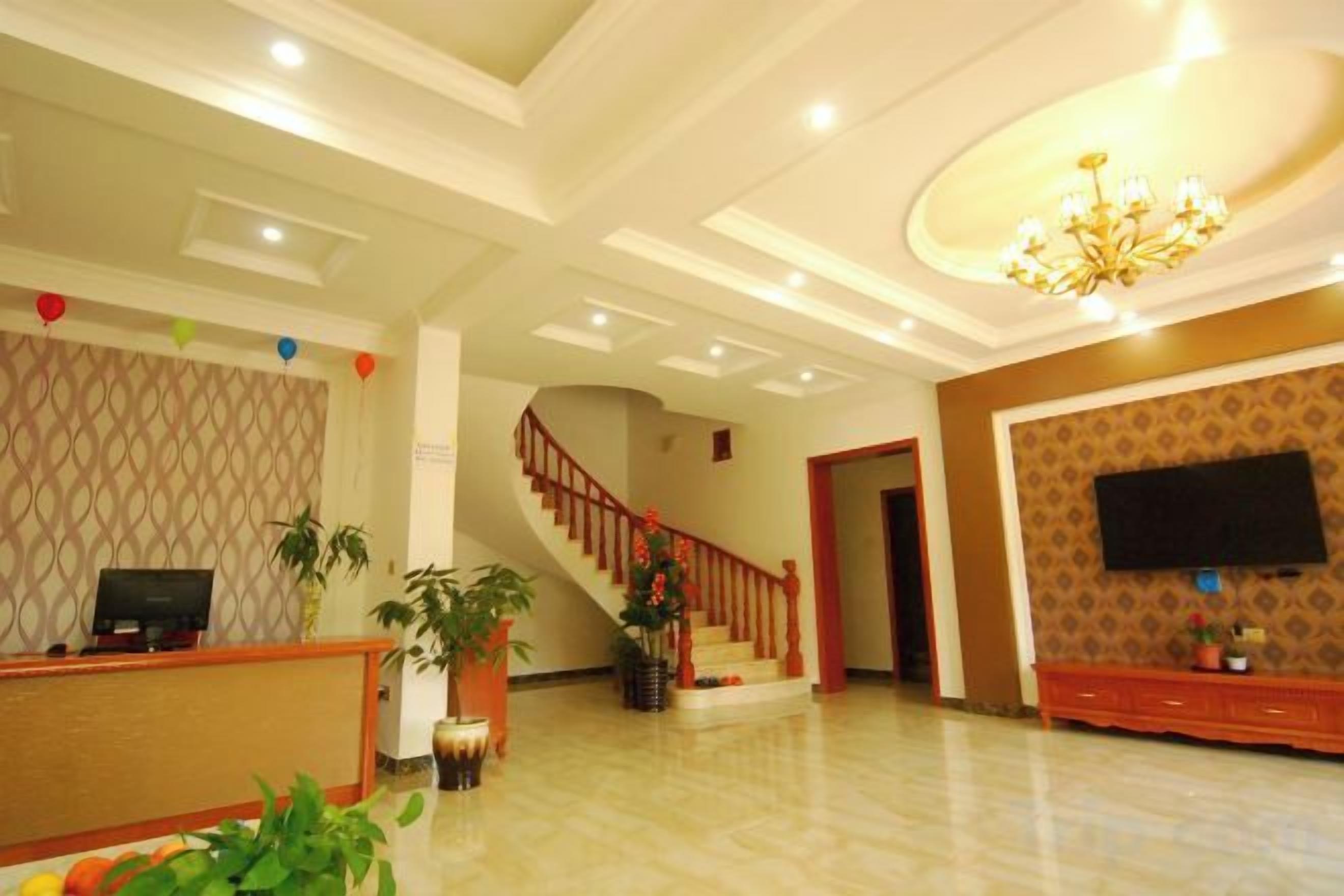 Un vestíbulo de hotel con una recepción, una escalera, un televisor de pantalla plana en la pared y un candelabro.