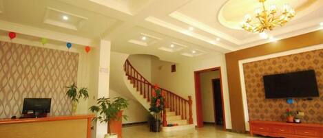 A hotel lobby with a reception desk, a staircase, a flat-screen TV mounted on the wall, and a chandelier.