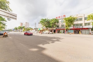 A street view with a red car driving on the road, buildings with storefronts, and trees lining the street. - Xianhong Business Hotel (Wanning High Speed Railway Station) (Wanning)