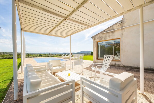 A covered outdoor seating area with white furniture, a pool, and a view of the landscape.