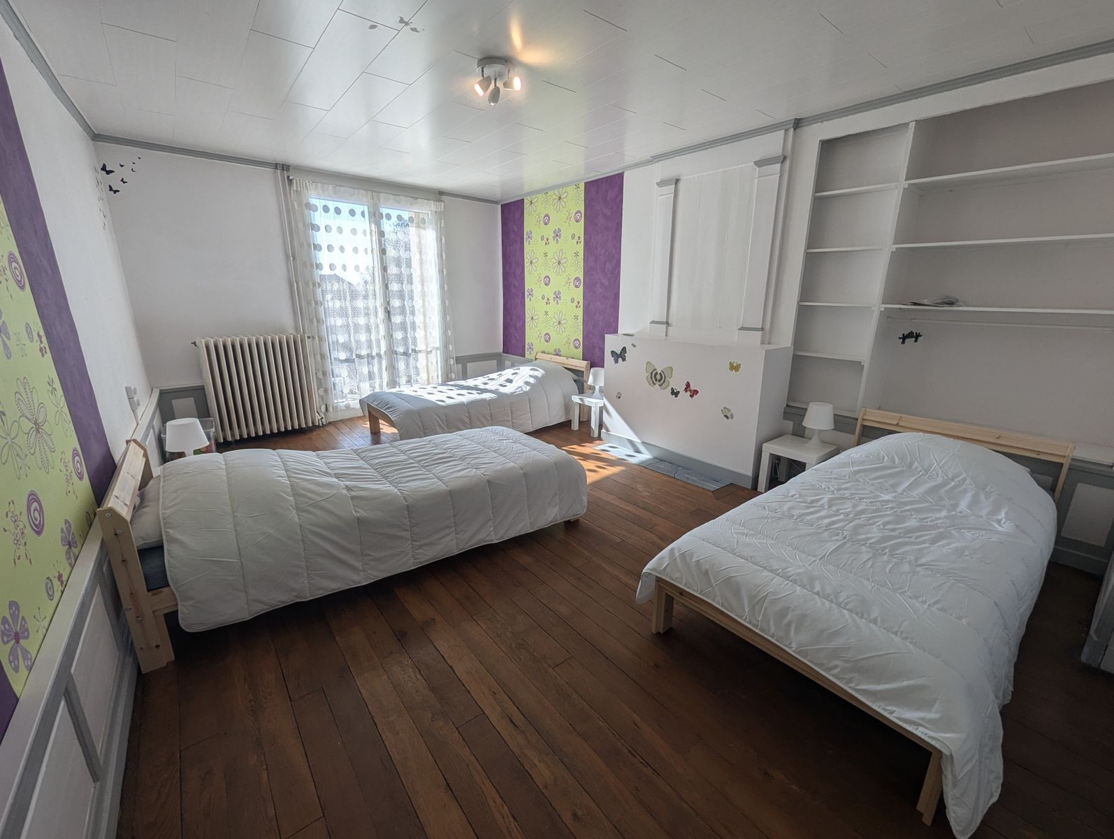 Habitación con dos camas individuales, un escritorio y una silla. Cuenta con un radiador y una ventana con cortinas.