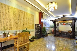 A lobby with a traditional Chinese calligraphy wall, a wooden reception desk, a wooden chair, a wooden gazebo, and potted plants.