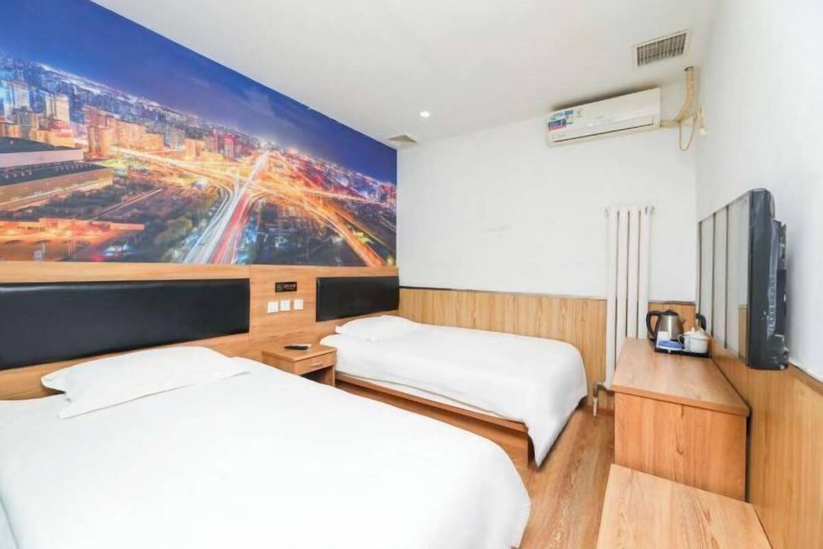 Habitación de hotel con dos camas, un mural de paisaje urbano en la pared, un equipo de aire acondicionado, un radiador y una mesita con un hervidor.