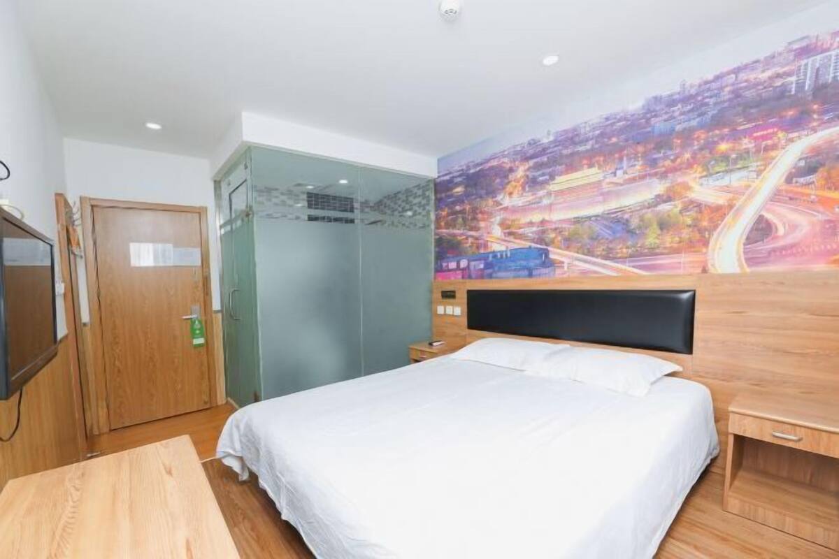 Una habitación de hotel con una cama, cabecera de madera, puerta de cristal y un mural con un paisaje urbano en la pared.