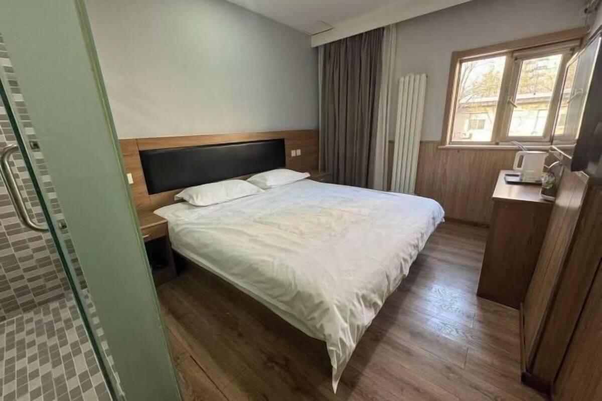 Una habitación de hotel con piso de madera, una cama con sábanas blancas, una mesita de noche, una ventana con cortinas y un escritorio de madera con una silla.