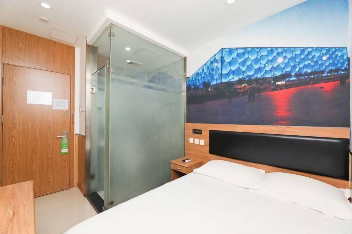 Habitación de hotel con cama, puerta de madera, cabina de ducha de vidrio y un amplio ventanal con vistas a un edificio de fachada redondeada.