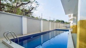 Una piscina rettangolare con bordo piastrellato, circondata da un muro basso e un edificio giallo.