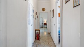 A hallway with white walls, a tiled floor, and framed pictures. There is a small table with a chair and a red bag.