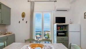 A dining area with a table, chairs, a bowl, a television, a bookshelf, and a view of the sea.