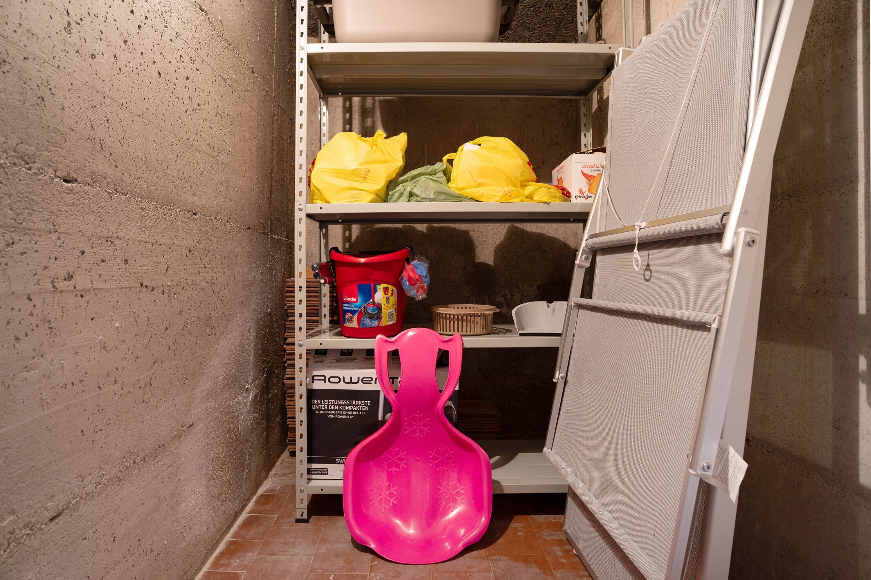 Un espace de rangement avec des étagères, une chaise en plastique rose et divers objets, notamment des sacs et une boîte.