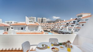 Un balcon avec une table dressée pour le petit-déjeuner, des chaises blanches et une vue sur des immeubles aux toits recouverts de tuiles rouges.