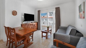 Un espace de vie compact comprenant un coin repas, un canapé, un téléviseur et un balcon agrémenté de rideaux.