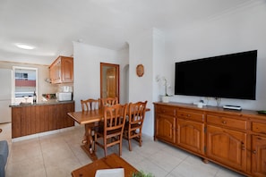 Une cuisine moderne avec des meubles en bois, une table à manger avec des chaises, un téléviseur posé sur un meuble et un petit îlot central équipé d’un four à micro-ondes et d’un grille-pain.