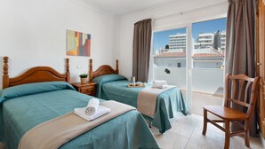 Une chambre d’hôtel avec deux lits, une chaise en bois, une petite table avec une plante et une vue sur les bâtiments, visible à travers une porte coulissante.