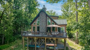 Une maison à deux étages avec une cheminée en pierre, une terrasse en bois et de grandes fenêtres.