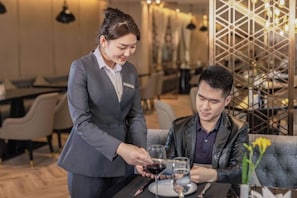 A person in a grey suit serving wine to another person seated at a table in a modern dining area with a geometric patterned wall and ambient lighting. - Tianhe Hotel (Guangzhou)
