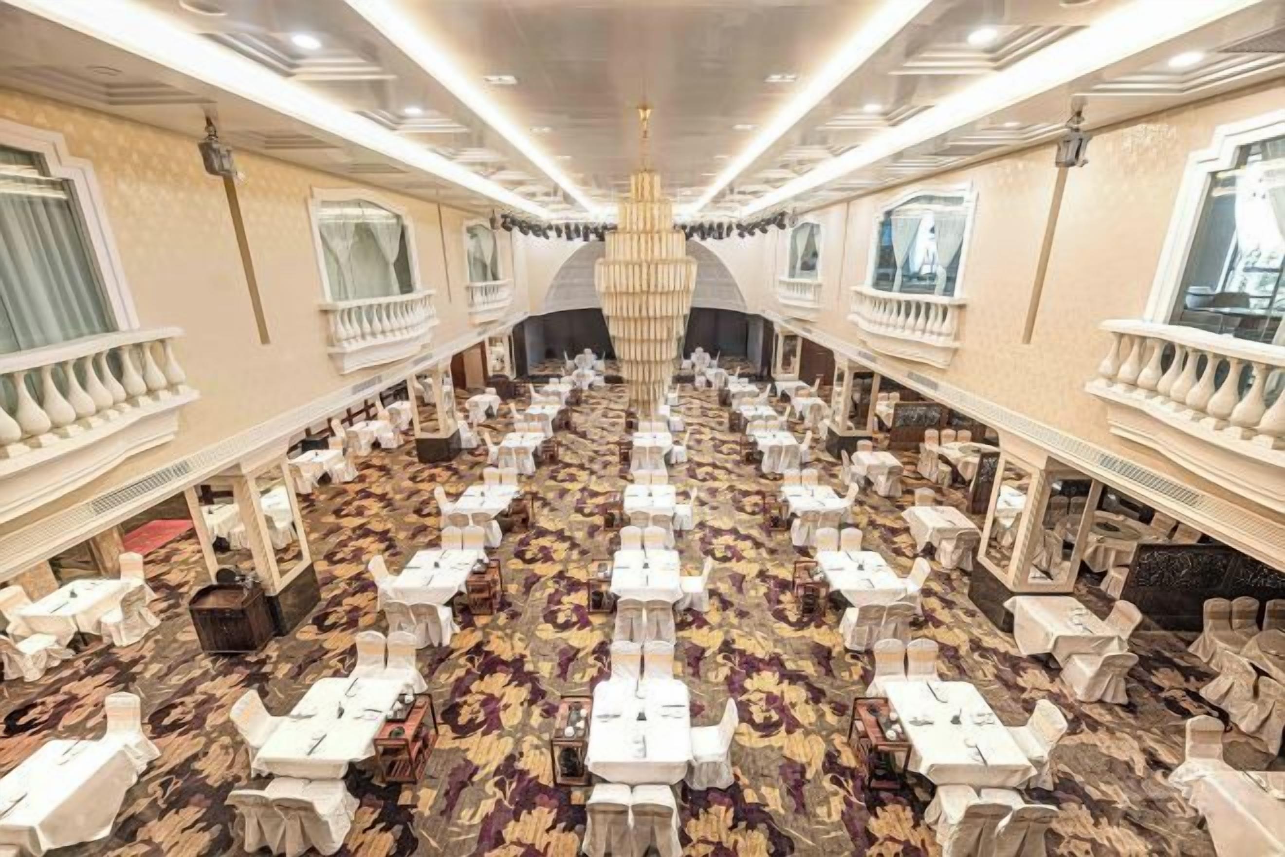Une salle de banquet avec de nombreuses tables disposées en rangées, un lustre central et un sol recouvert de moquette.