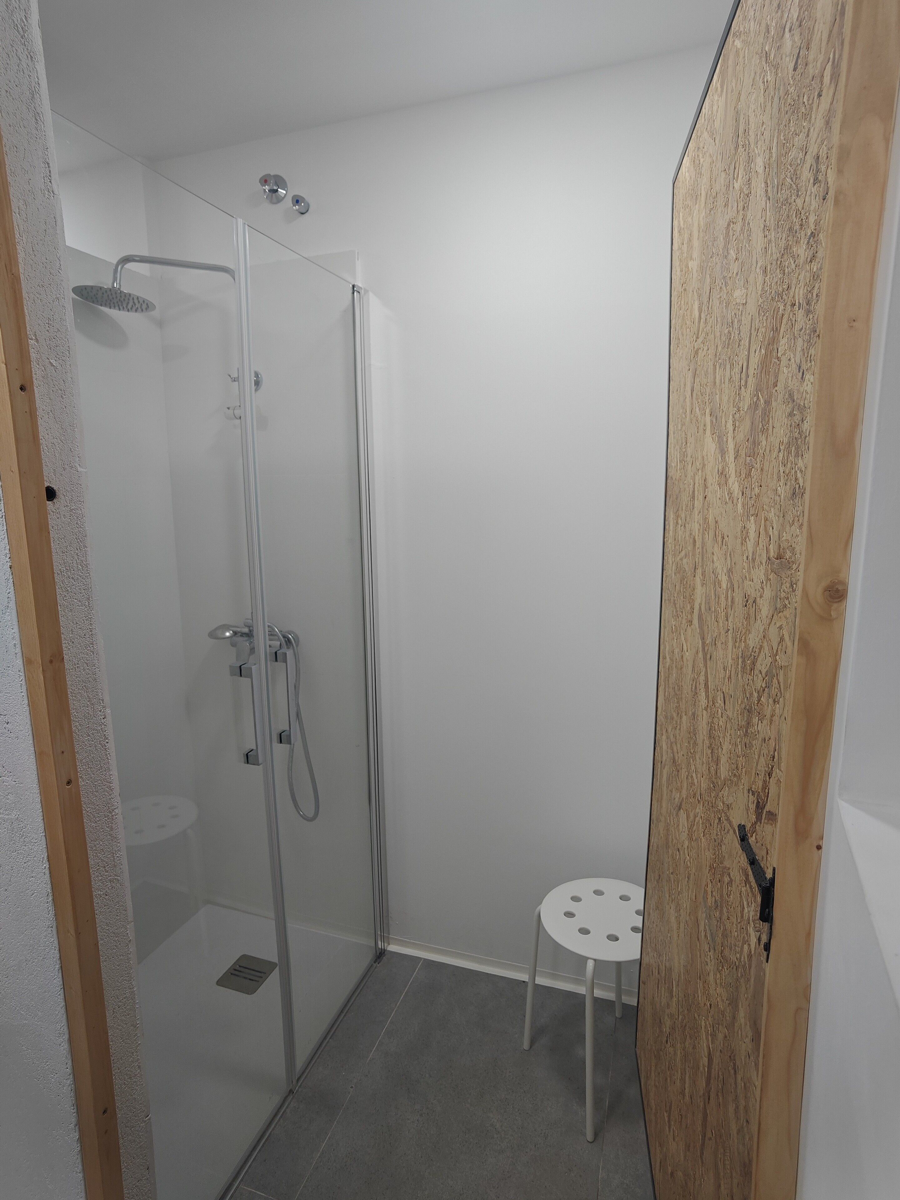 Un baño pequeño y moderno con cabina de ducha de vidrio, un taburete y una puerta de madera.