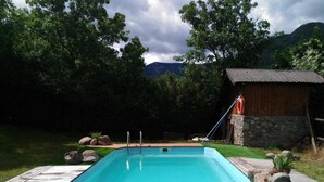 A rectangular swimming pool with a wooden deck, surrounded by rocks and grass, with a wooden cabin in the background. - Cal Caló – Colònies Cadí is a mountain farmhouse for peaceful groups in Cortariu, La Cerdanya. (Bellver De Cerdanya)