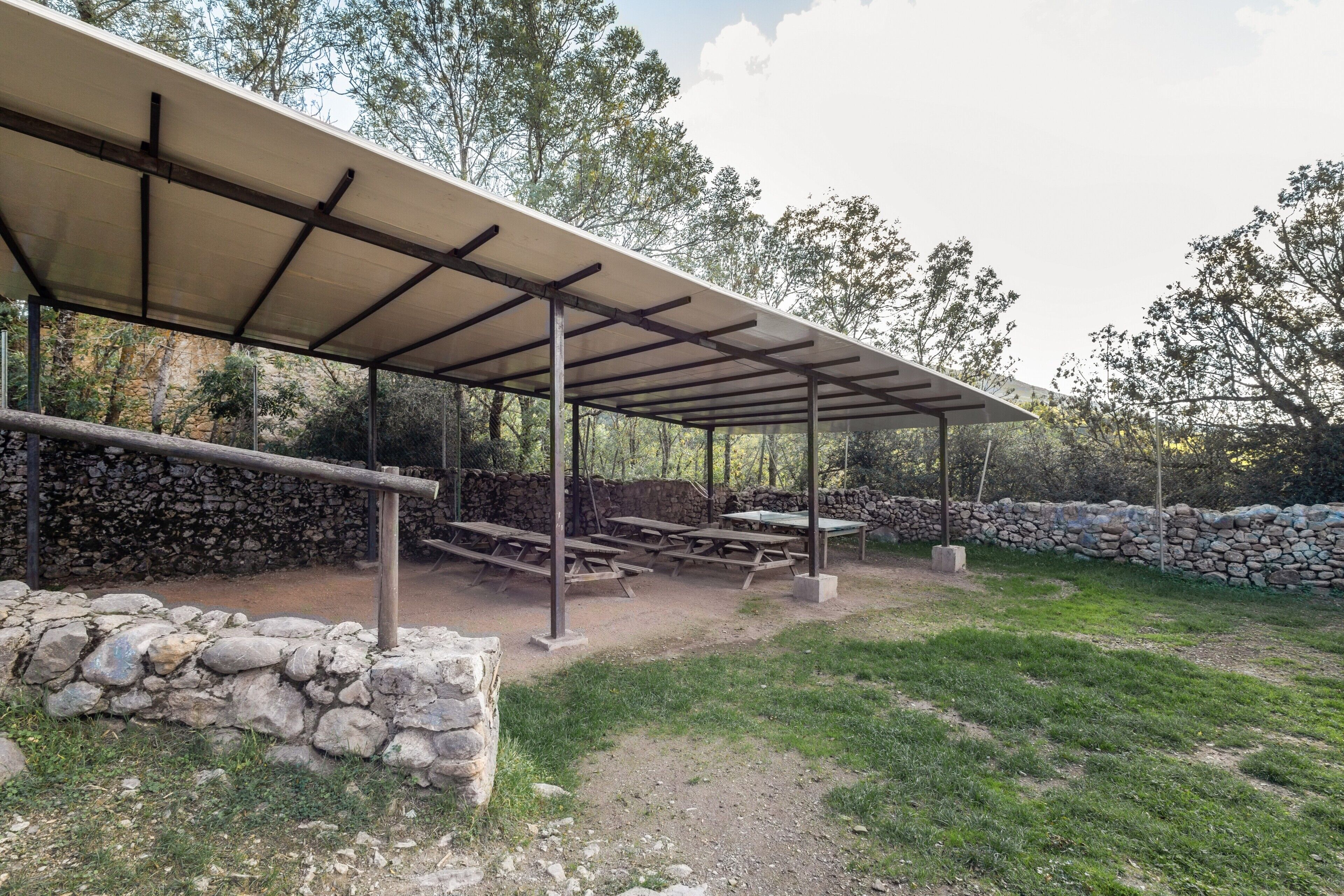 Un espace pique-nique abrité, doté de tables et de bancs, entouré de murs en pierre et d’arbres.