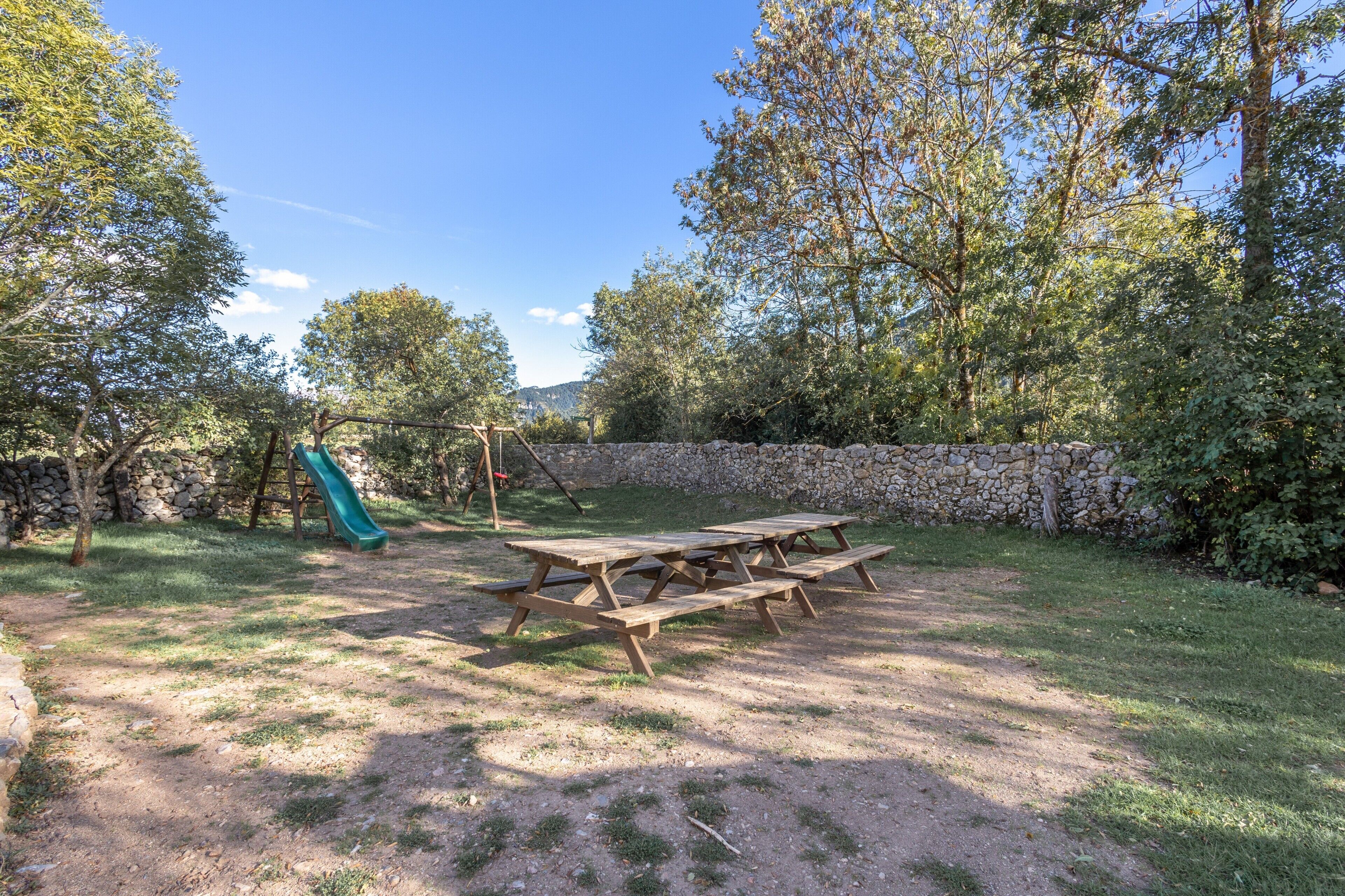 Una mesa de picnic en un área verde con un muro de piedra y árboles al fondo.