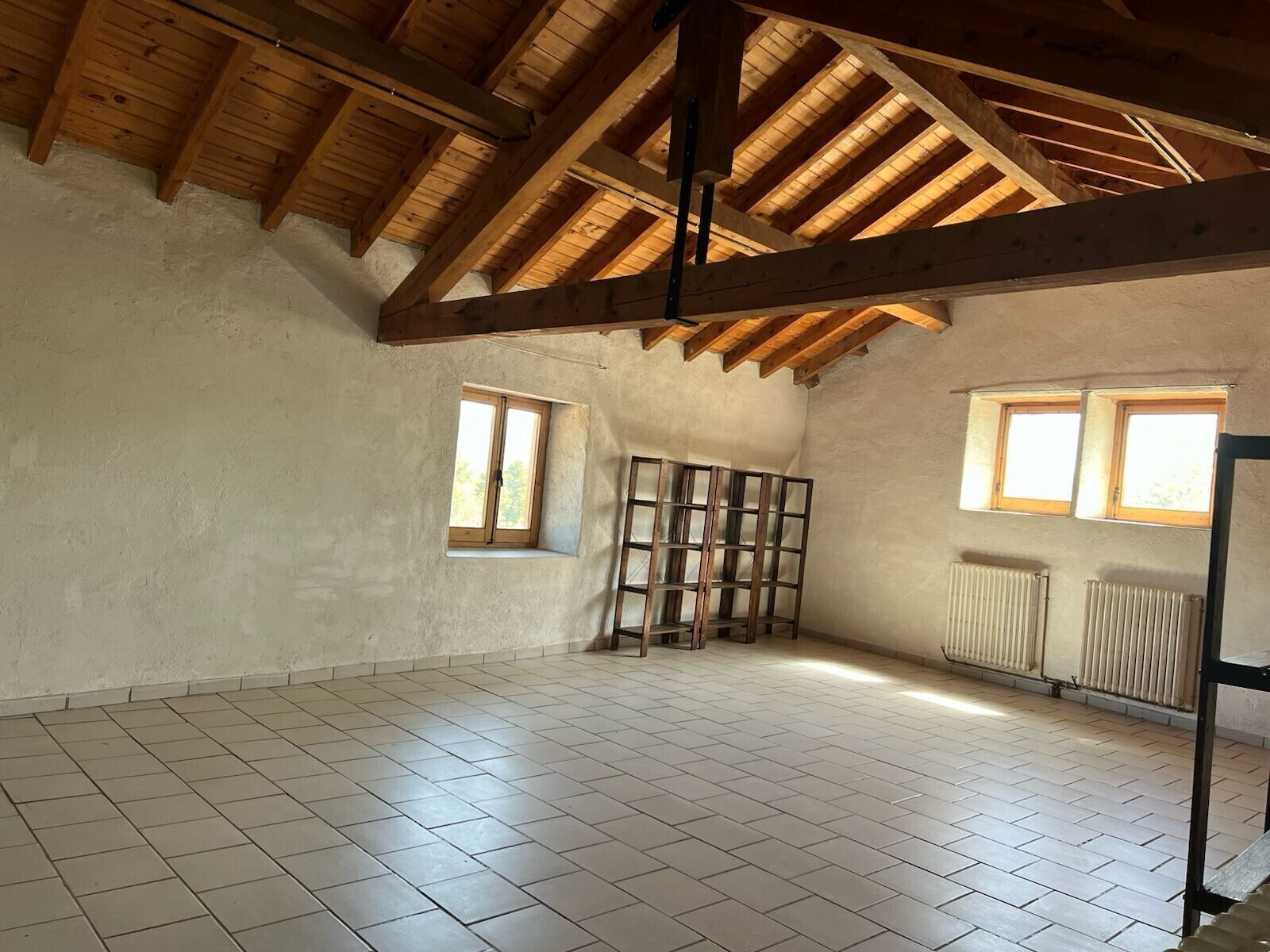 Una habitación vacía con techo de madera inclinado, suelo de baldosas y dos ventanas.