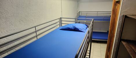 A small, narrow room with bunk beds, a wooden shelf, and a white wall.