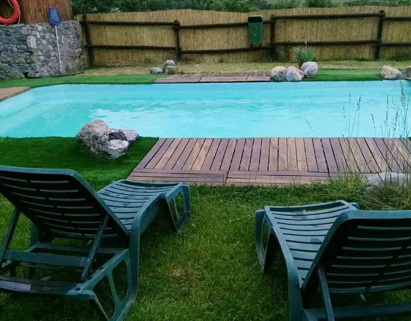 Une piscine avec une terrasse en bois, deux chaises longues et un mur de pierre en arrière-plan.