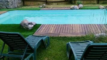 Une piscine avec une terrasse en bois, deux chaises longues et un mur de pierre en arrière-plan.