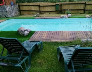 Een zwembad met een houten terras, twee ligstoelen en een stenen muur op de achtergrond.