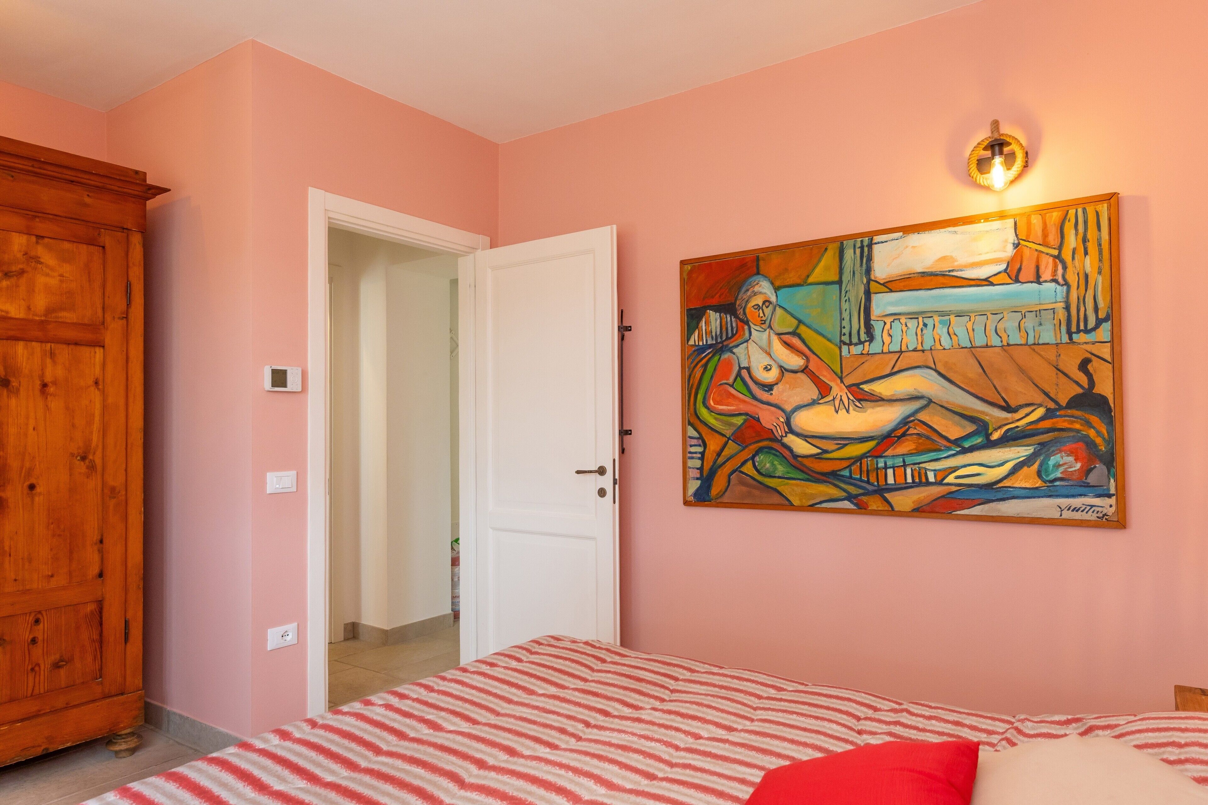 A bedroom with a bed, a wooden wardrobe, a door leading to another room, and a painting on the wall.