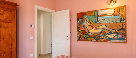 A bedroom with a bed, a wooden wardrobe, a door leading to another room, and a painting on the wall.