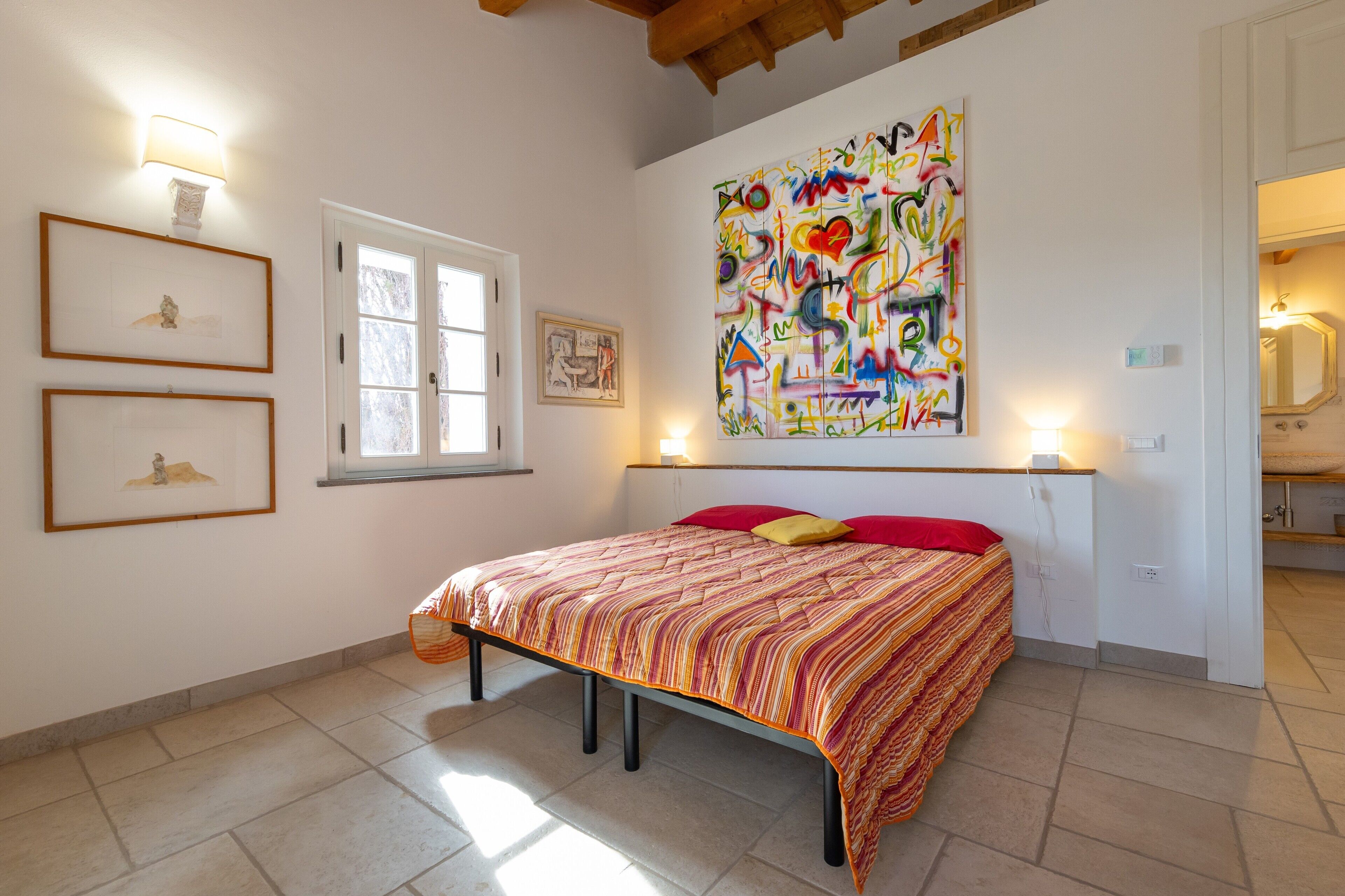 Een slaapkamer met een bed, een raam, een muur met ingelijste kunst en een kleurrijk abstract schilderij.