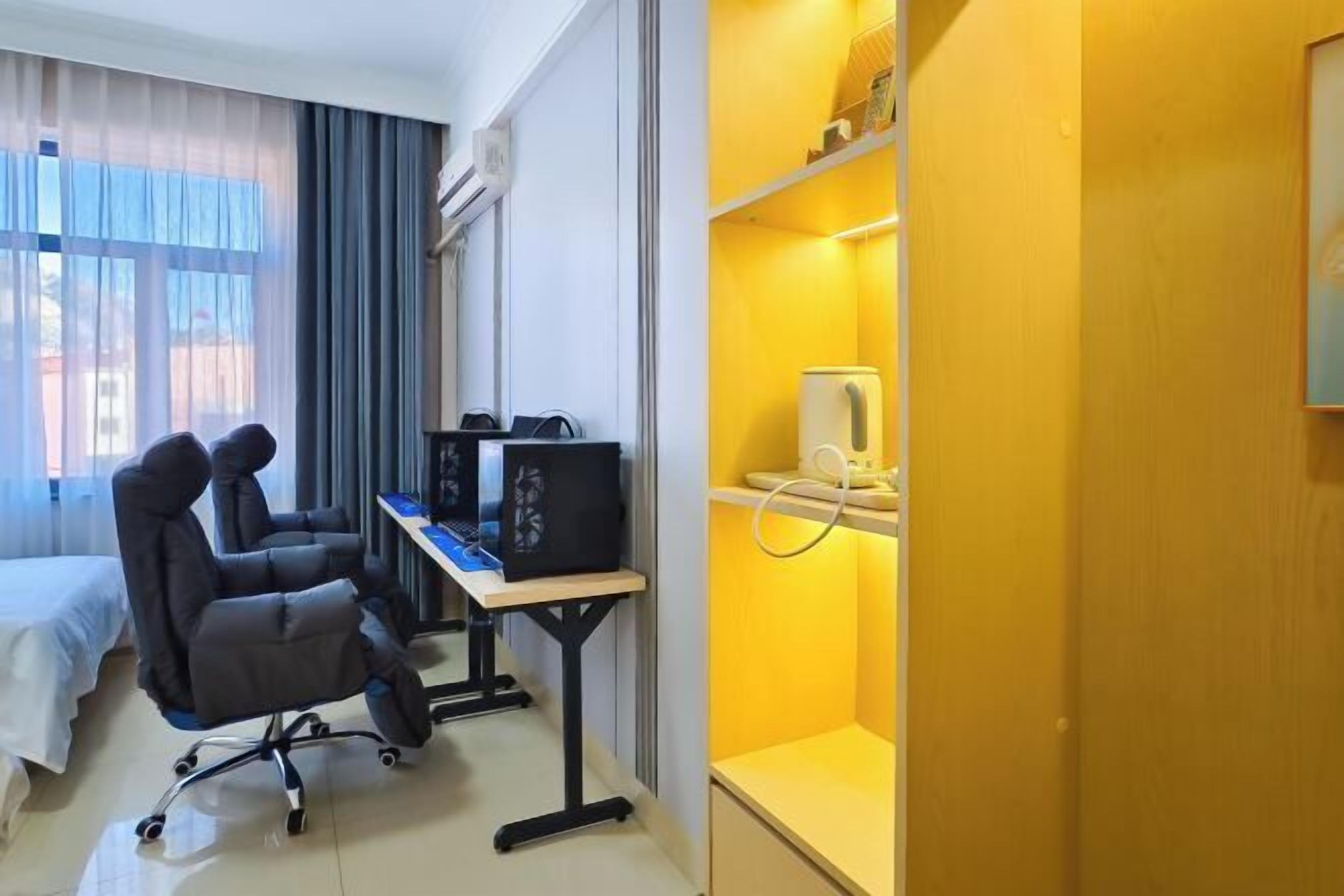 A hotel room with a bed, a desk with a computer, a chair, and a shelf with a lamp.