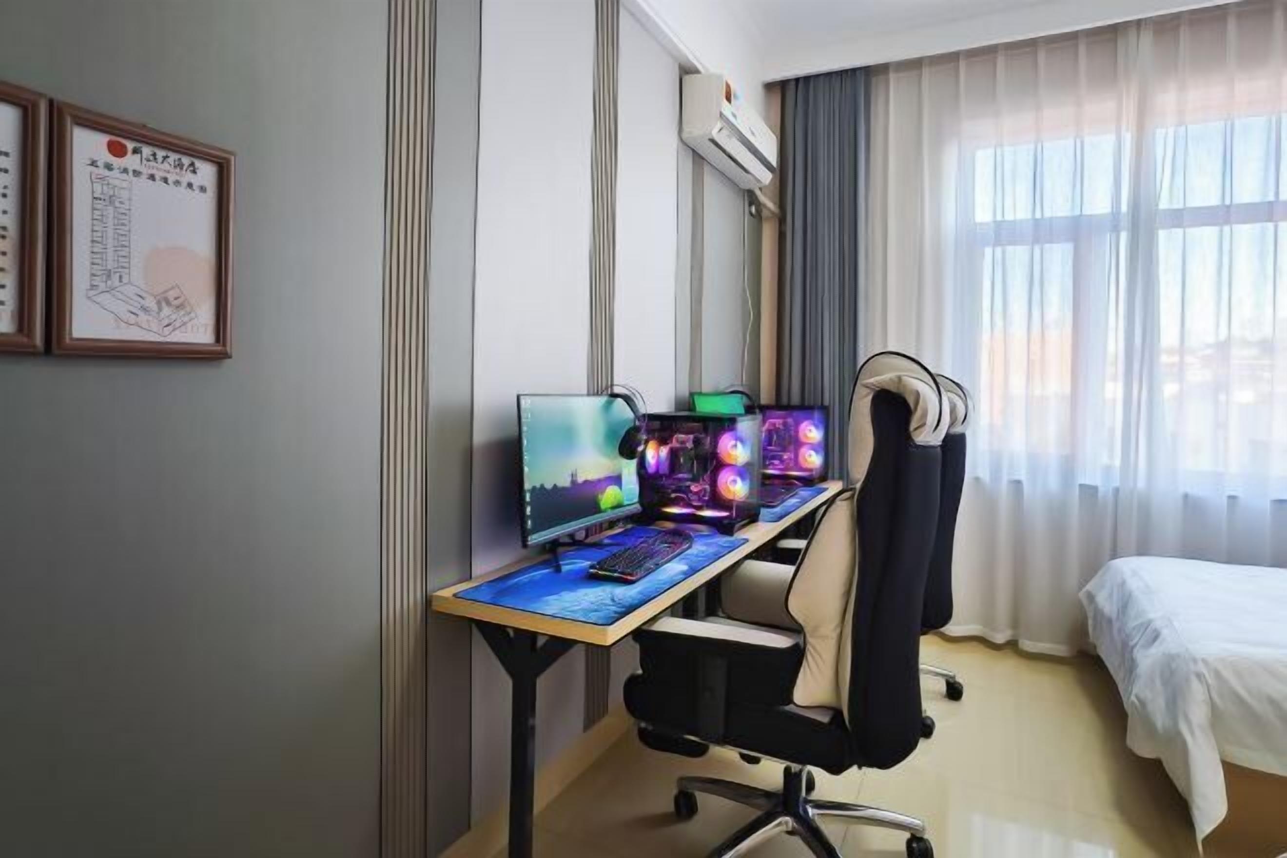 A hotel room with a desk, two monitors, a keyboard, a chair, a bed, and a window with curtains.