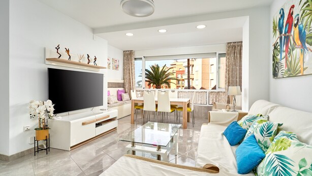 A modern living room with a flat-screen TV, a glass coffee table, a sofa with patterned cushions, and a dining area with chairs and a table.