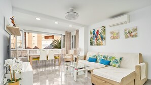 A modern living room with a glass coffee table, beige sofa, and tropical-themed wall art.