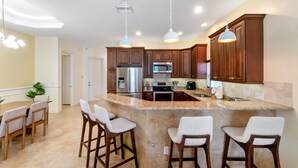 Una cocina moderna con encimeras de granito, electrodomésticos de acero inoxidable y armarios de madera. Hay tres taburetes de bar y una mesa de comedor con sillas.
