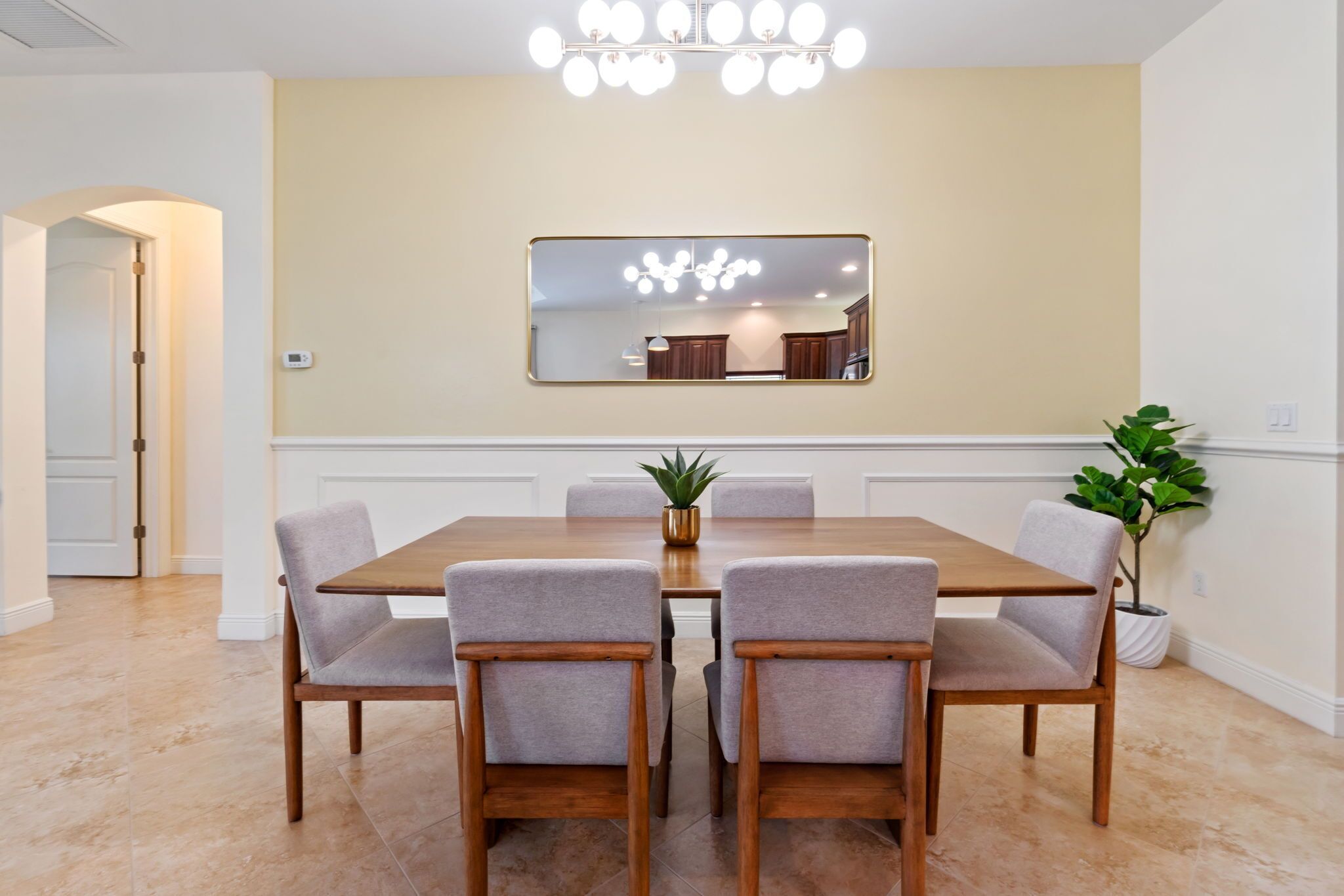 A dining area with a rectangular table, six grey upholstered chairs, a potted plant, and a wall-mounted mirror reflecting the room.
