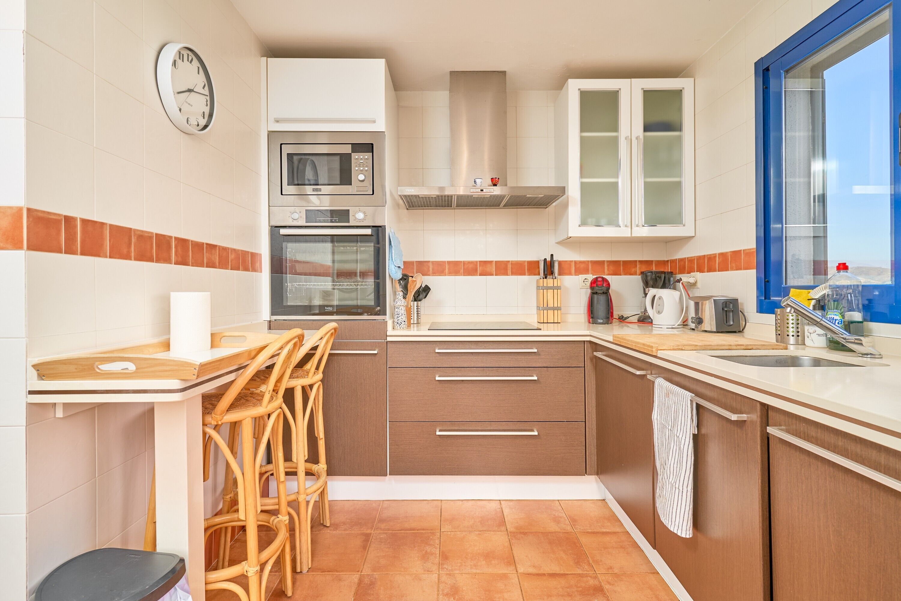 A kitchen with a microwave, oven, and cabinets, a window, and a clock on the wall.