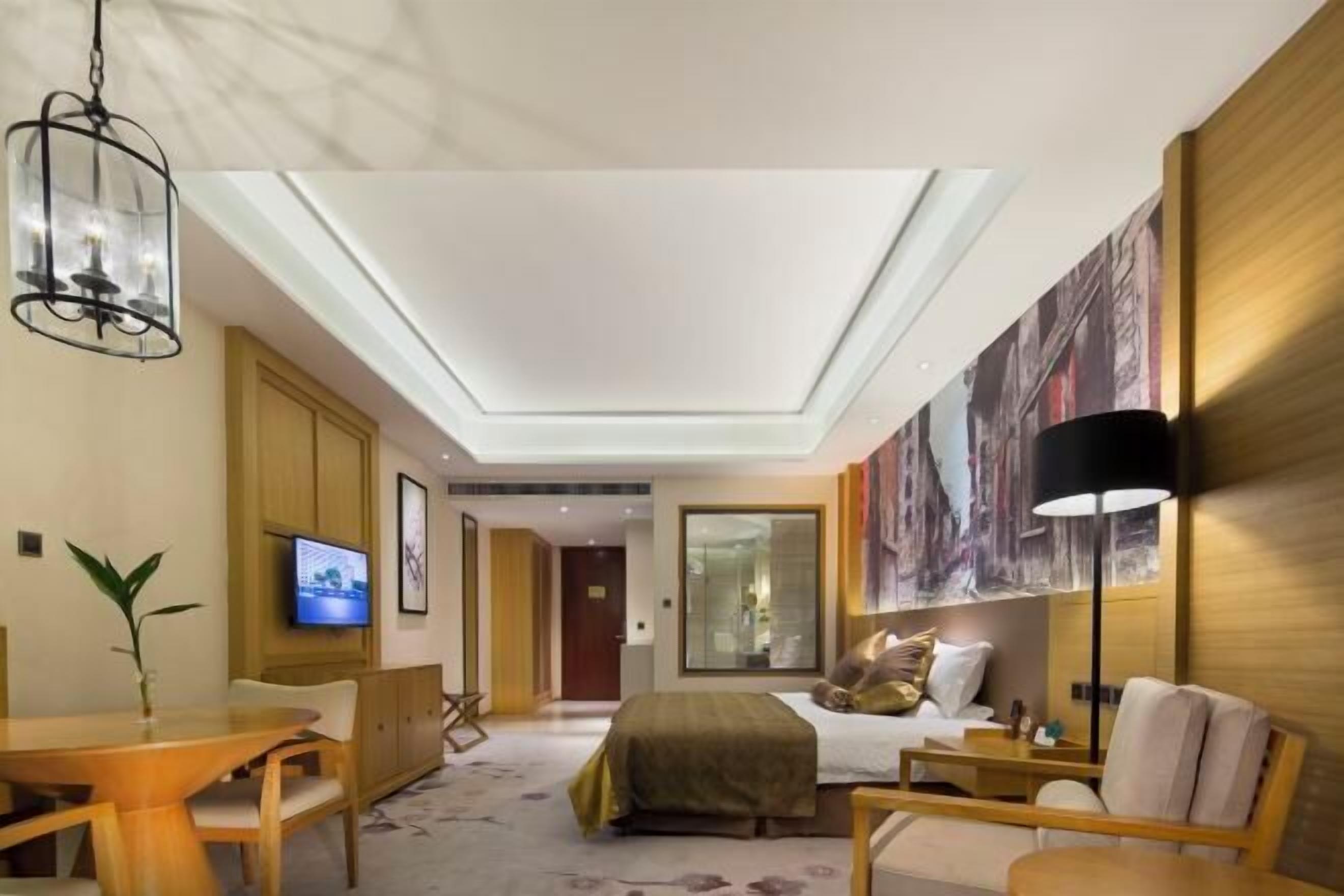A modern hotel room with a large bed, a desk, a chair, a TV, and a ceiling light.