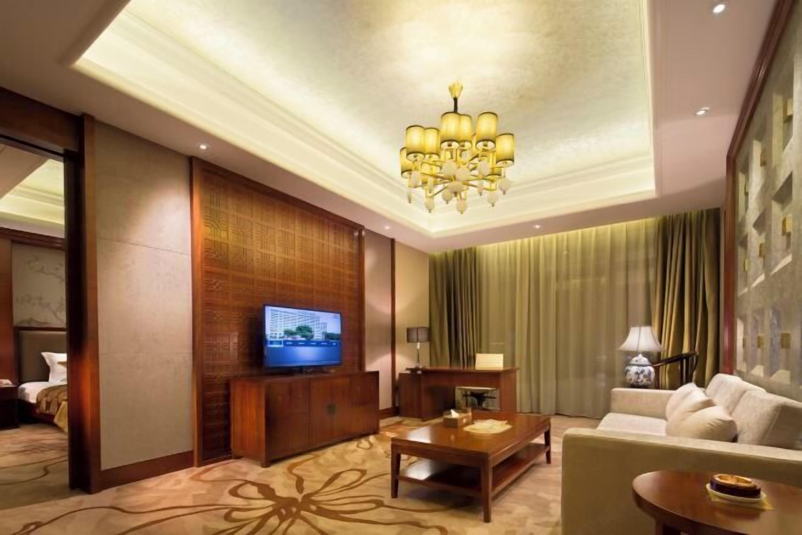 Una habitación de hotel con televisión, mueble de entretenimiento de madera, sofá, mesa de centro y candelabro.