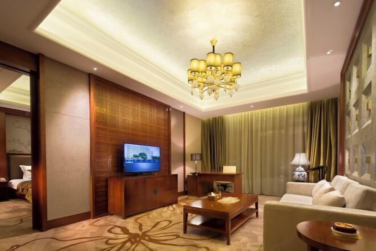 A hotel room with a television, a wooden entertainment unit, a sofa, a coffee table, and a chandelier.