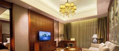 A hotel room with a television, a wooden entertainment unit, a sofa, a coffee table, and a chandelier.