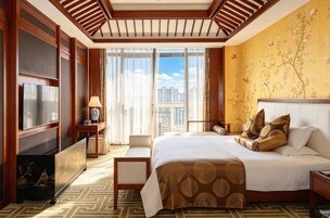 Habitación de hotel con una cama grande, televisión, escritorio con lámpara y vista a la ciudad a través de amplios ventanales.