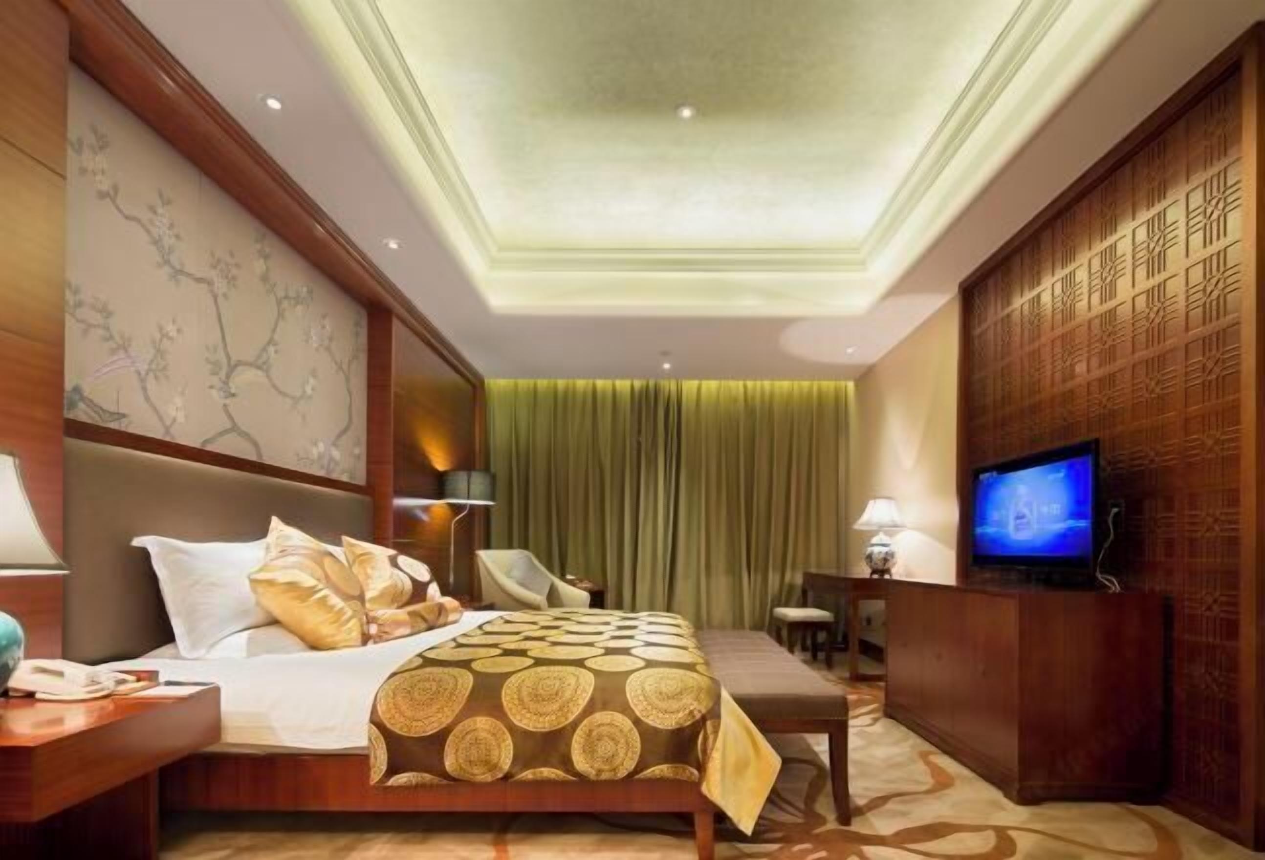 A hotel room with a bed, bedside table, lamp, TV, and a desk with a chair.