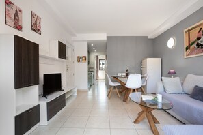 A modern living room with a flat-screen TV, a glass coffee table, a sofa, and a dining area with chairs and a table.